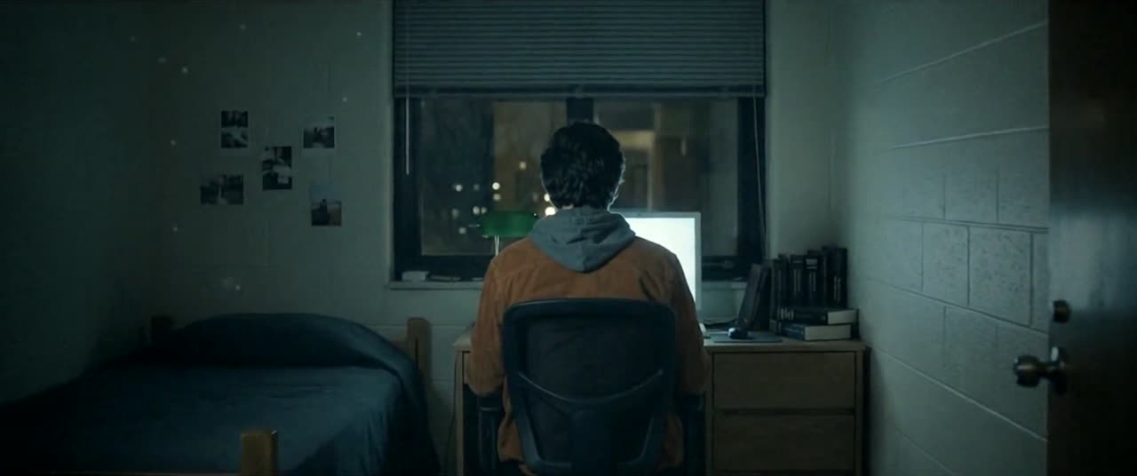 Night. A figure in a rust jacket sits at a dorm desk in the glow of a monitor, back to camera