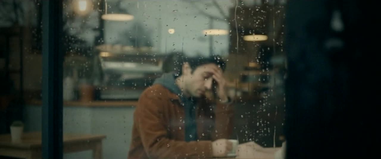 Eli seen through a rain-streaked cafe window, hand to his forehead